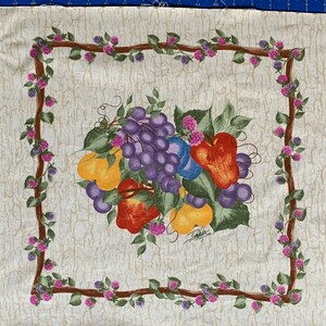 Op de afbeelding: Een stoffen paneel met een stilleven van fruit in het midden, waaronder druiven, peren en paprika's. Het ontwerp is omlijst door een bruine rand met wijnranken en roze bloemen. De achtergrond is een getextureerd off-white.
