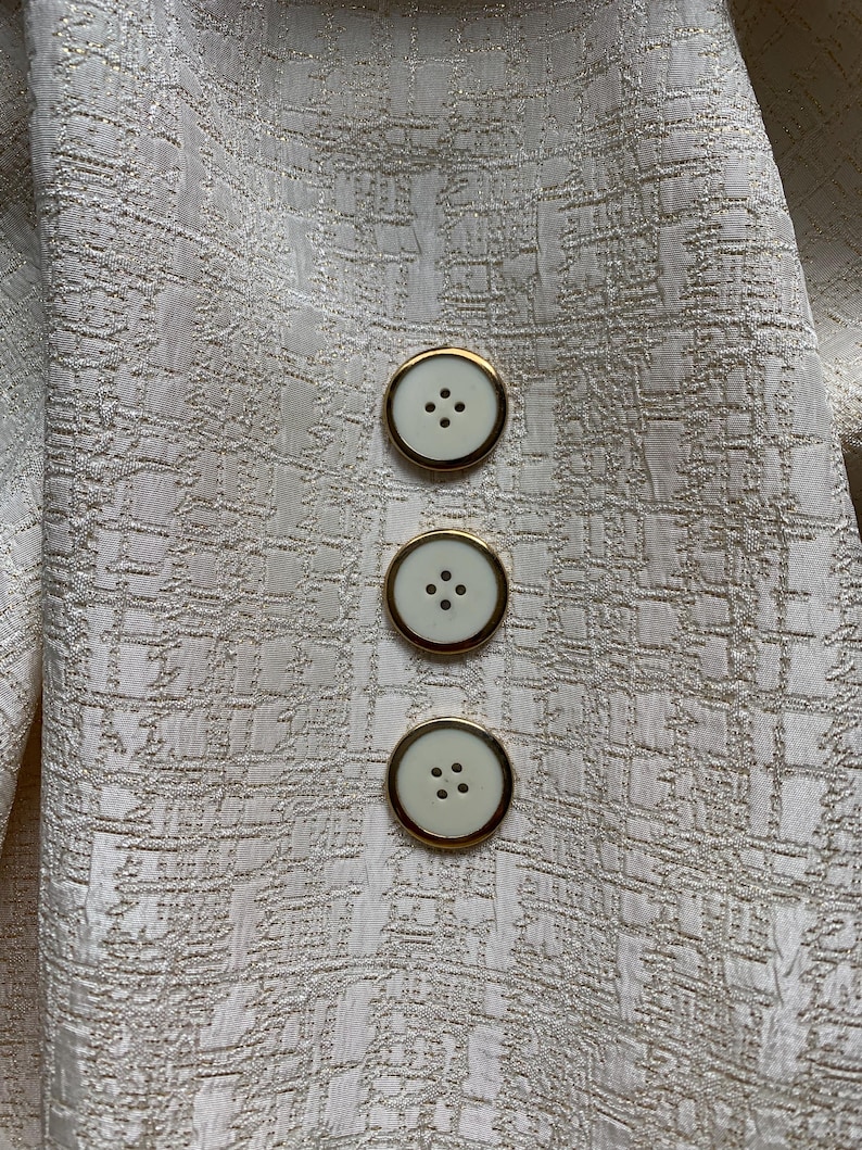 Puede incluir: Una tela de color crema con un patr&oacute;n texturizado en forma de cuadr&iacute;cula con hilos dorados met&aacute;licos. Tres botones circulares est&aacute;n alineados verticalmente, cada uno con un centro crema liso, cuatro agujeros peque&ntilde;os y un borde de metal dorado.