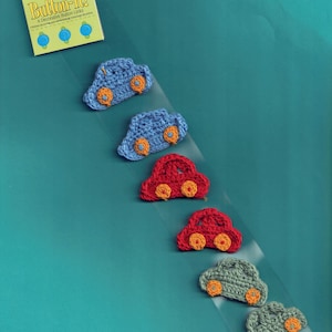 Pode incluir: Um pacote de botões decorativos "Button-it!" com seis botões de crochê em forma de carro nas cores azul, vermelho e verde, cada um com rodas laranja. A embalagem é amarela e azul, com o texto "Button-it!" e "Daisy Kingdom".