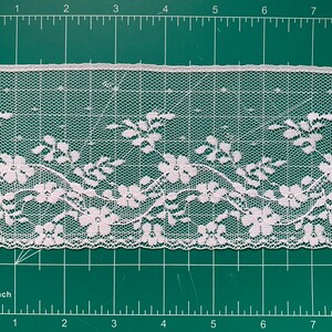 May include: A strip of white floral lace trim featuring a repeating pattern of five-petaled flowers and delicate vines. The lace is positioned against a green cutting mat with a visible ruler, showing a width of approximately 1.5 inches.