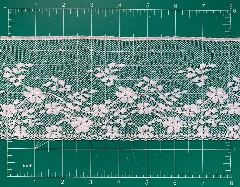 May include: A strip of white floral lace trim featuring a repeating pattern of five-petaled flowers and delicate vines. The lace is positioned against a green cutting mat with a visible ruler, showing a width of approximately 1.5 inches.