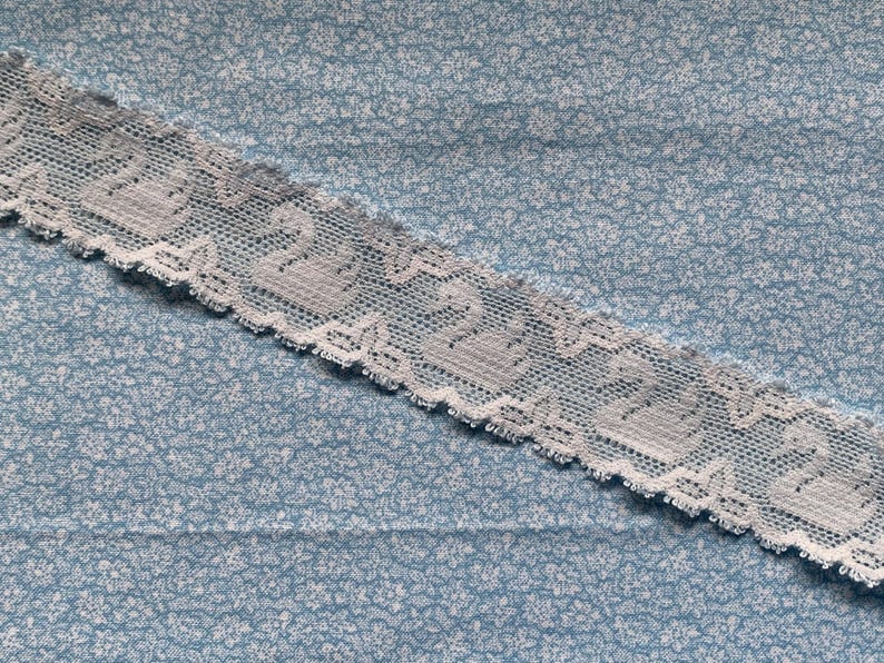 May include: A strip of white lace trim featuring a repeating pattern of swans, positioned diagonally across a light blue fabric background that is covered in a dense, small-scale white floral print. The lace has a scalloped edge detail.