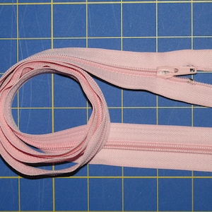 以下が含まれることがあります： 金属製の滑り止めと引き手のあるピンクのジッパー。ジッパーは巻き上げられ、青いグリッド状の表面に平らに置かれています。