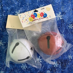 Puede incluir: Dos cascabeles redondos, uno blanco y otro rojo, cada uno con una ranura negra en forma de cruz. Est&aacute;n empaquetados en bolsas de pl&aacute;stico transparente con una tarjeta de cabecera Nicole Carnival of Crafts con gr&aacute;ficos de confeti.