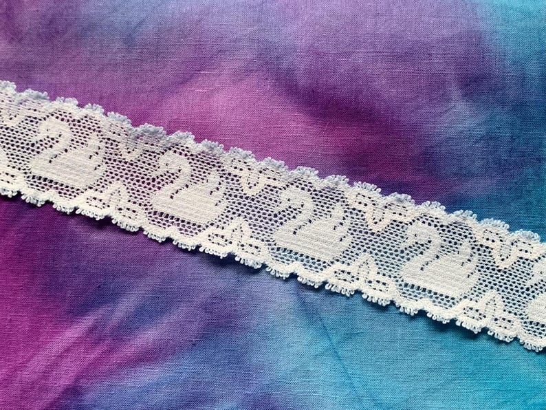 May include: A strip of white lace trim featuring a repeating pattern of swans. The lace has a scalloped edge and a delicate mesh texture, showcasing a series of swan motifs oriented in the same direction across the length of the fabric trim.