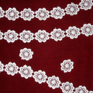 Puede incluir: Borde de encaje blanco con diseño floral sobre fondo rojo. El encaje está dispuesto en un patrón de tres filas, siendo la fila superior la más ancha y la fila inferior la más estrecha.