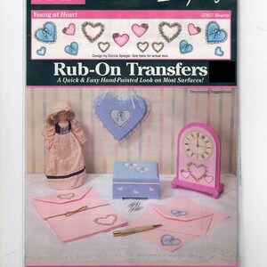 May include: A package of rub-on transfers featuring a heart design. The package shows decorating suggestions for a clock, a box, envelopes, and a heart-shaped plaque. The text on the package reads "PLAID Accents On Everything Young at Heart 57057 Hearts Rub-On Transfers A Quick & Easy Hand-Painted Look on Most Surfaces! Great for Wood, Paper, Plastic, Metal, Ceramics & More! Contains 1 sheet with 15 designs and applicator. Samples shown may use more than 1 transfer sheet."