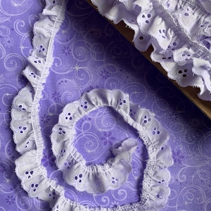 May include: A roll of white eyelet lace with a scalloped edge and small circular cutouts. The lace is a soft white color and is displayed on a purple fabric with a floral and swirl pattern. The lace is approximately 1 inch wide.