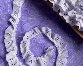 May include: A roll of white eyelet lace with a scalloped edge and small circular cutouts. The lace is a soft white colour and is displayed on a purple fabric with a floral and swirl pattern. The lace is approximately 2.5 cm wide.