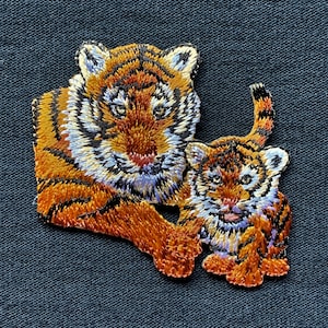 May include: Embroidered patch of a mother tiger and her cub. The mother tiger is orange and black with white stripes. The cub is smaller and has a more orange color. Both tigers have black noses and white whiskers.