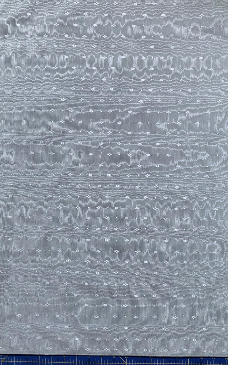 May include: A close-up of a gray fabric with a textured pattern. The fabric features horizontal wavy lines and small diamond-shaped white accents, creating a subtle, elegant design. The overall appearance is soft and flowing.