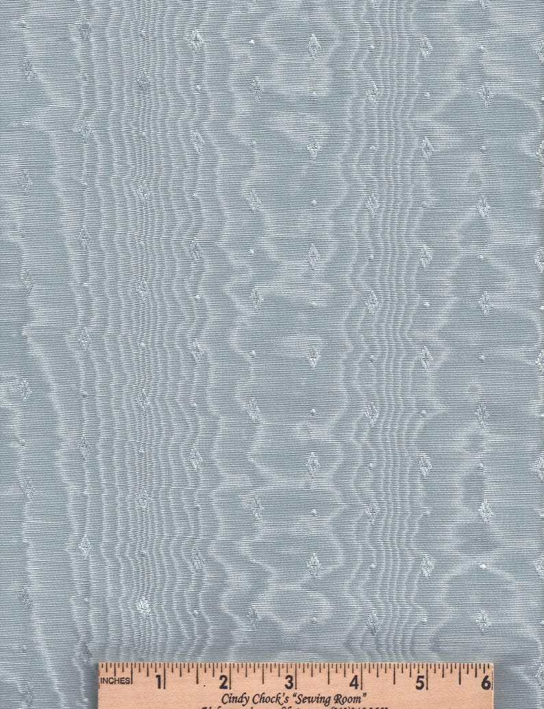 May include: Light blue fabric with a subtle, rippled texture and small diamond-shaped accents. The fabric has a delicate, shimmering appearance. A wooden ruler with inch measurements is at the bottom of the image.