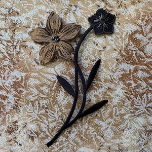 May include: A black and gold fabric flower applique with beaded accents. The flower is attached to a black fabric stem with leaves.