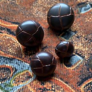 May include: Four dark brown, spherical buttons with a cracked, geometric design. The buttons vary in size and are arranged on a patterned, textured surface with shades of brown, orange, and blue.