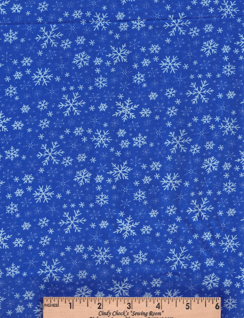 May include: A blue fabric featuring a repeating pattern of white snowflakes in various sizes. A wooden ruler is positioned at the bottom, showing a scale of 6 inches with the text Cindy Chock's Sewing Room printed on the ruler.