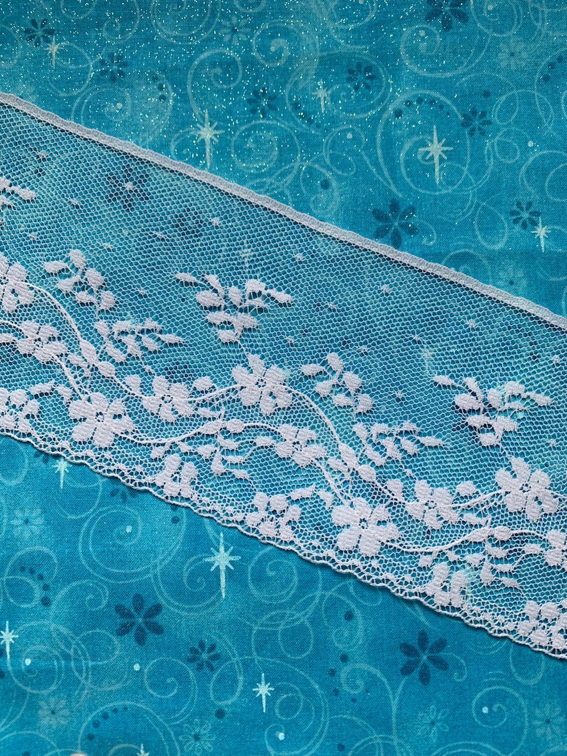 May include: A strip of white lace trim featuring a delicate floral pattern with scalloped edges. The lace has a fine mesh background and is positioned diagonally across a bright turquoise fabric background decorated with small stars and swirls.