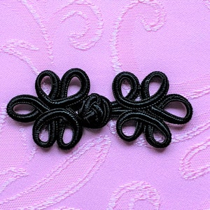 May include: Black Chinese knot closure with a decorative design. The knot is made of a thick, black cord and has a traditional knot pattern.