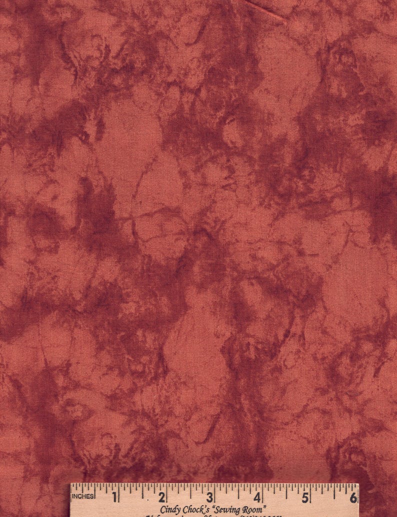May include: A piece of fabric with a mottled, marbled pattern in shades of deep red and terracotta. A wooden ruler is positioned at the bottom edge, showing a scale of 6 inches, with the text Cindy Chock's Sewing Room printed on it.