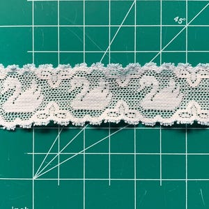 May include: A strip of white lace trim featuring a repeating pattern of swans, positioned over a green cutting mat with a visible grid and inch markings, showing the lace width is approximately 3.2 centimetres across the horizontal axis.