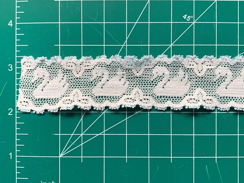 May include: A strip of white lace trim featuring a repeating pattern of swans, positioned over a green cutting mat with a visible grid and inch markings, showing the lace width is approximately 3.2 centimetres across the horizontal axis.