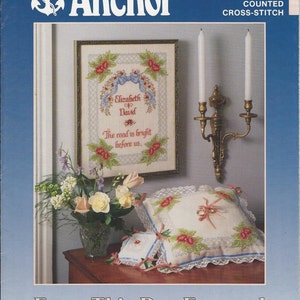 May include: The cover of an "Anchor" counted cross-stitch kit. The image features a framed cross-stitch with floral and ribbon designs, a vase of flowers, and decorative pillows. Text reads "Elizabeth & David" and "The road is bright before us."