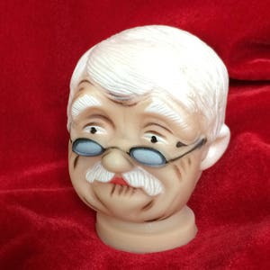 Puede incluir: Figura de cerámica con la cabeza de un anciano con pelo blanco, bigote blanco y gafas con montura azul. La figura tiene un tono de piel neutro y está sobre un fondo rojo. La cabeza está montada sobre una base cilíndrica corta.