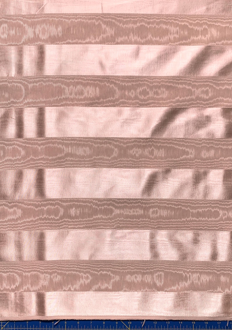 May include: Close-up of a fabric with a striped pattern. The fabric has a light pink background with horizontal stripes in a slightly darker shade of pink. The stripes have a textured, wood-grain-like appearance, creating a subtle contrast.