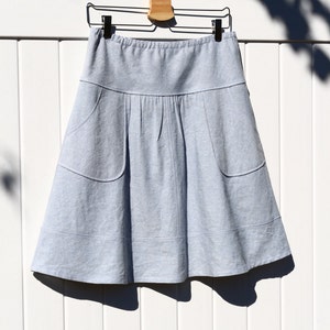 May include: A light blue skirt with two pockets hanging on a wooden hanger against a white background.