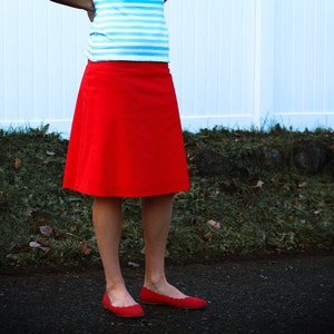 May include: A woman wearing a red skirt and a blue and white striped shirt. She is standing in front of a white fence and green grass.