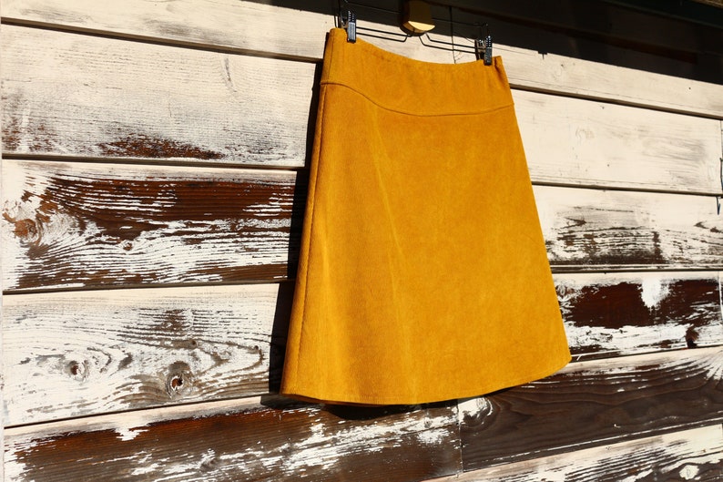 May include: A mustard yellow A-line skirt hanging on a wooden hanger against a white and brown wood background.