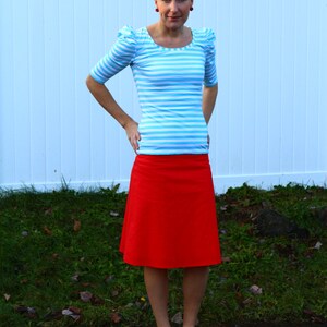 May include: A woman wearing a blue and white striped shirt with puffed sleeves and a red skirt. She is standing in front of a white fence with green grass in the background.