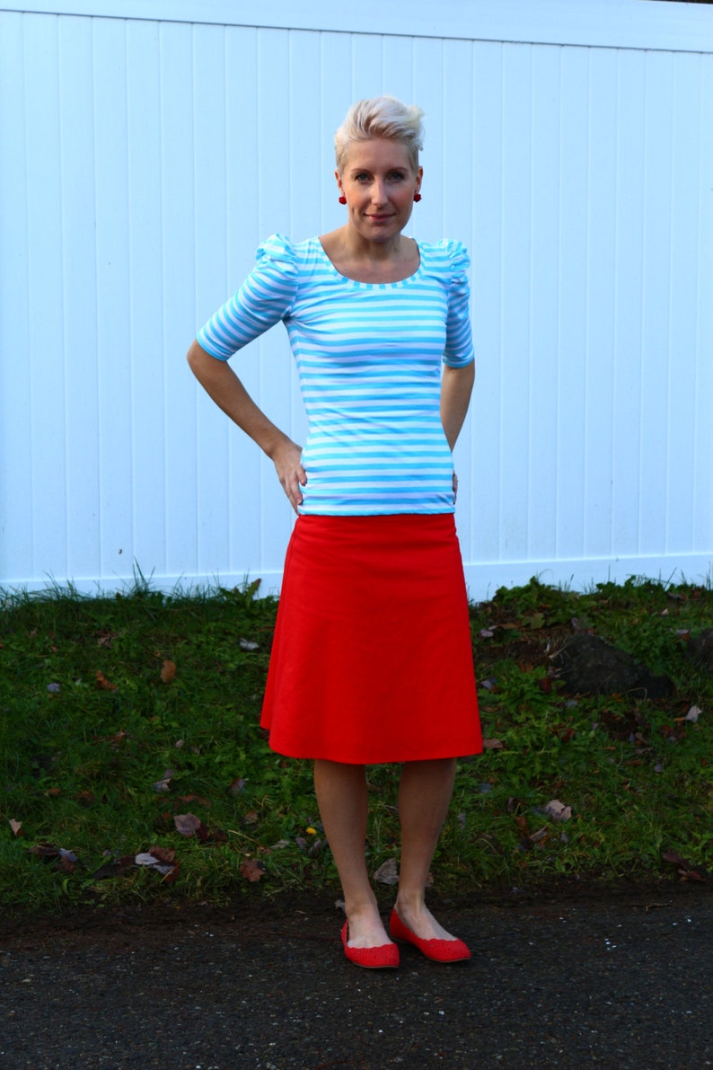 May include: A woman wearing a blue and white striped shirt with puffed sleeves and a red skirt. She is standing in front of a white fence with green grass in the background.