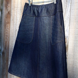 May include: A dark blue denim skirt with a waistband and two pockets. The skirt is hanging on a wooden hanger.