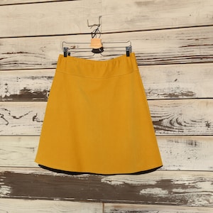 May include: A mustard yellow A-line skirt hanging on a wooden hanger against a whitewashed wood background.
