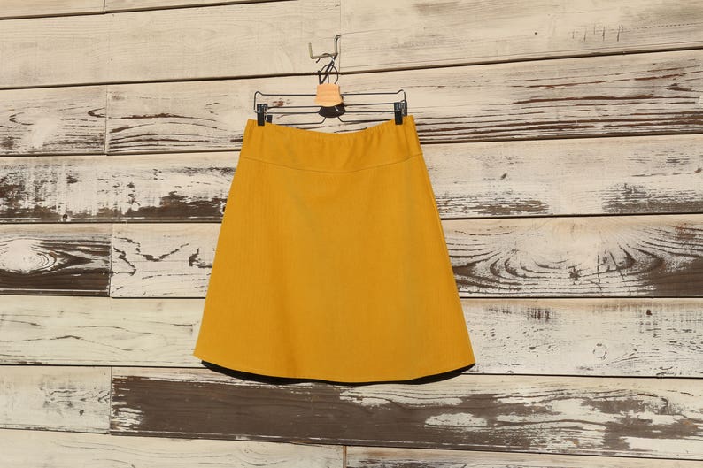 May include: A mustard yellow A-line skirt hanging on a wooden hanger against a whitewashed wood background.