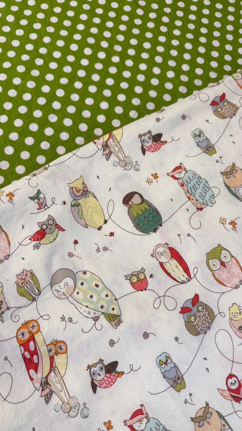 May include: White fabric with a pattern of colorful owls in various poses, connected by curved lines. The owls have different colors and patterns. A section of green fabric with white polka dots is in the upper left corner.