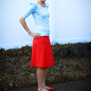 May include: A woman wearing a blue and white striped shirt with a red skirt and red shoes. She is standing in front of a white fence.
