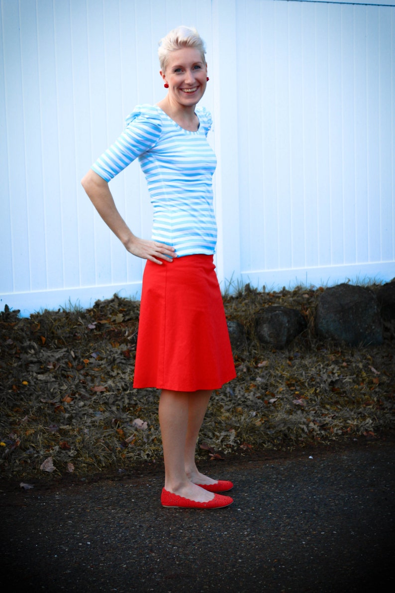 May include: A woman wearing a blue and white striped shirt with a red skirt and red shoes. She is standing in front of a white fence.