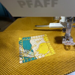 May include: A close-up of a sewing machine with a needle and thread. A small square piece of fabric with a floral pattern is being sewn onto a larger piece of yellow corduroy fabric.