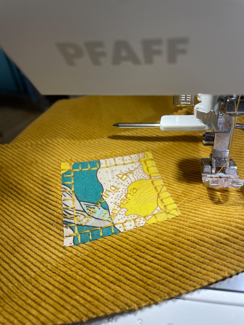 May include: A close-up of a sewing machine with a needle and thread. A small square piece of fabric with a floral pattern is being sewn onto a larger piece of yellow corduroy fabric.