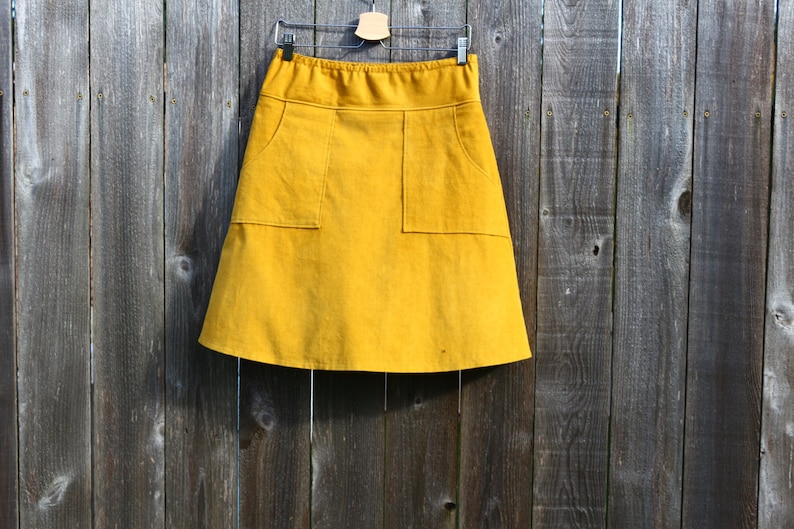 May include: A mustard yellow skirt with two large pockets on the front. The skirt is hanging on a wooden hanger against a wood panel background.