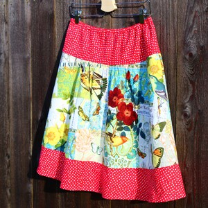 May include: A-line skirt with a red polka dot waistband and hem. The skirt features a patchwork design with floral, bird, and butterfly patterns in shades of yellow, blue, and green. The skirt is hanging on a wooden background.