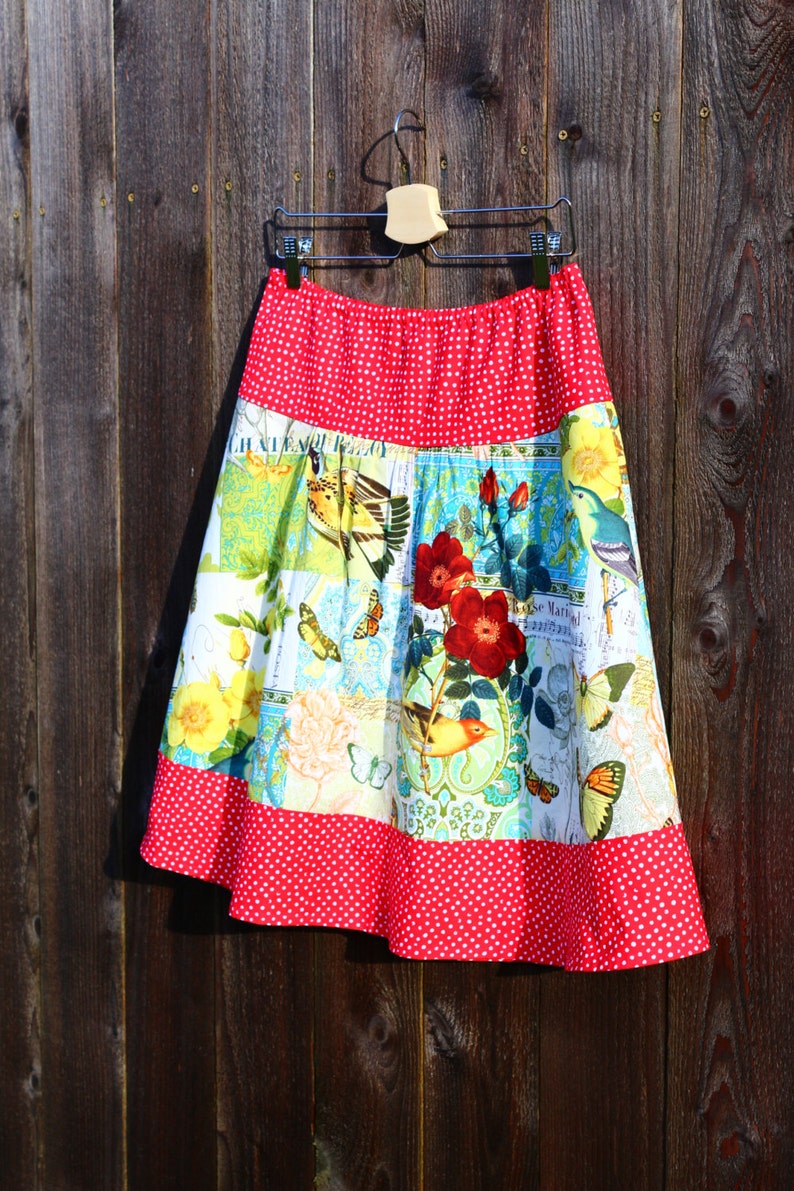 May include: A-line skirt with a red polka dot waistband and hem. The skirt features a patchwork design with floral, bird, and butterfly patterns in shades of yellow, blue, and green. The skirt is hanging on a wooden background.