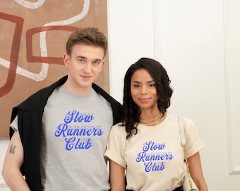 Puede incluir: Dos personas con camisetas grises y crema con el texto "Slow Runners Club" en letras azules.