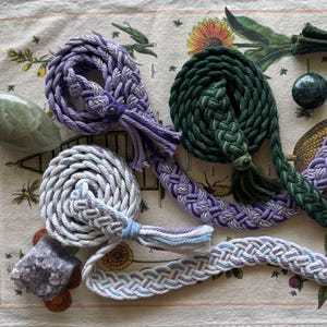 May include: An assortment of hand-braided rope items in various colors, including purple, green, and white. The ropes are arranged on a patterned fabric, with some coiled and others in elongated shapes. Decorative tassels and crystals are also present.