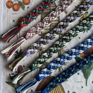 May include: Braided friendship bracelets in various colors, including green, purple, and blue, with decorative beads and tassels. The bracelets are displayed on a floral patterned fabric.
