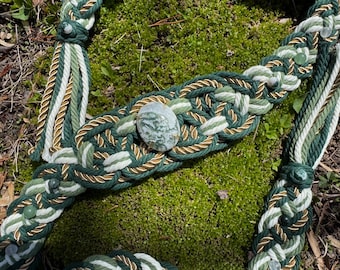 May include: A handcrafted, braided rope belt in shades of green, white, and gold. The belt features a central stone accent and decorative tassels. The design incorporates various shades of green, creating a natural, earthy aesthetic.