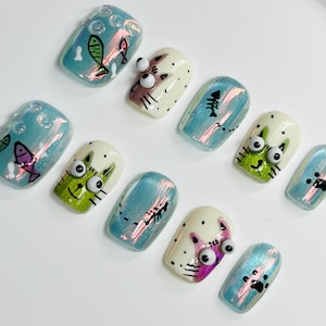 May include: Set of ten blue and white false fingernails with various cat and fish designs. The designs include a cat with googly eyes, a fish with a tail, and a paw print.