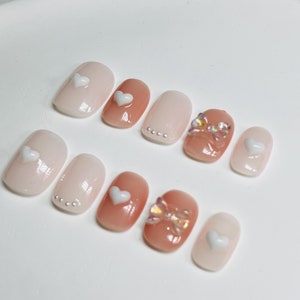 Puede incluir: Conjunto de diez uñas postizas de color rosa y blanco con diseños de corazón y lazo.