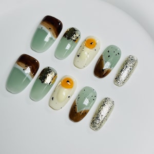 May include: Set of ten green and brown fake nails with a variety of designs, including a fried egg, gold leaf, and glitter.